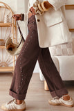 A person wearing brown wide-leg cuffed pants with silver stud detailing, styled with a cream blazer, beige sneakers, and a woven straw bag in a bright indoor setting.