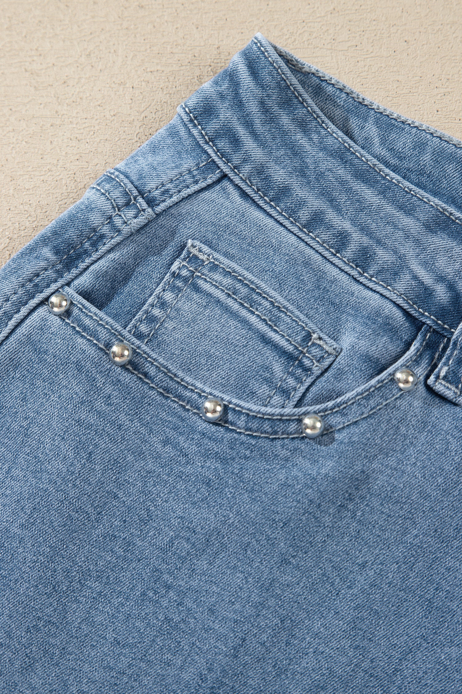 Close-up of the back pocket of light blue denim jeans decorated with five silver-toned pearl-like studs along the pocket edge, set against a neutral beige textured background.