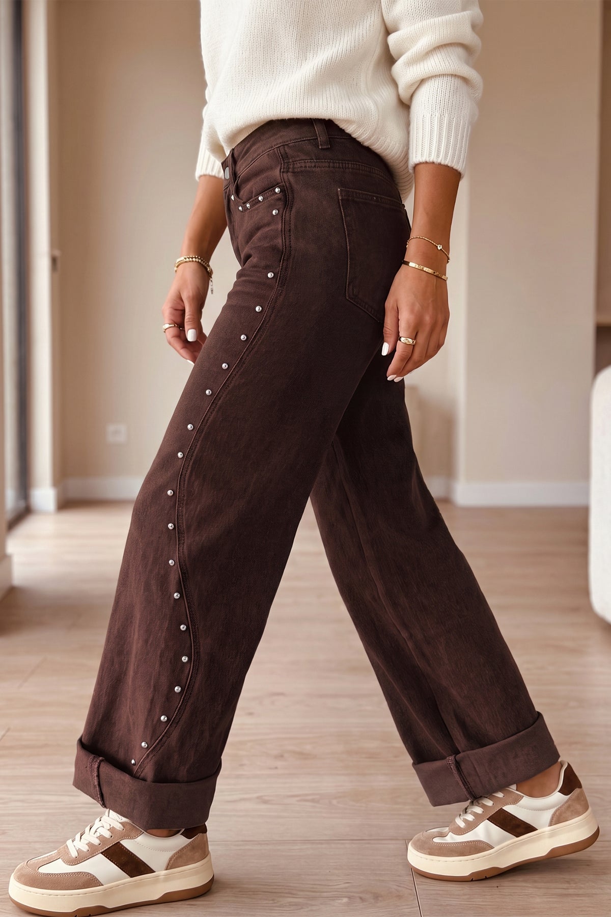 A person wearing dark brown wide-leg jeans with silver stud embellishments down the side, paired with a cream sweater and beige-and-white sneakers in an indoor room setting.