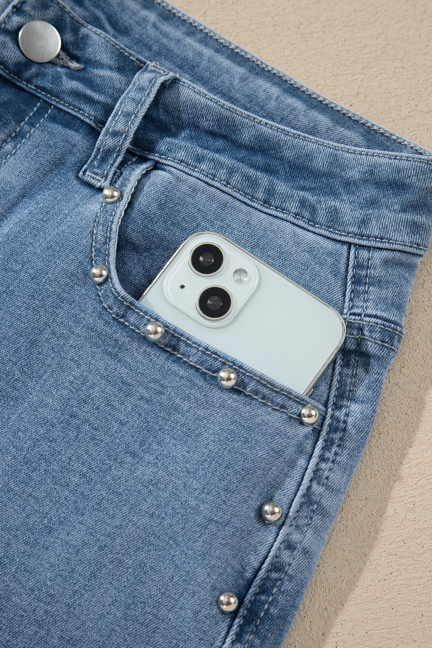 Close-up of a light blue denim jacket with silver stud embellishments on the pocket, showing a light blue smartphone tucked into the pocket.