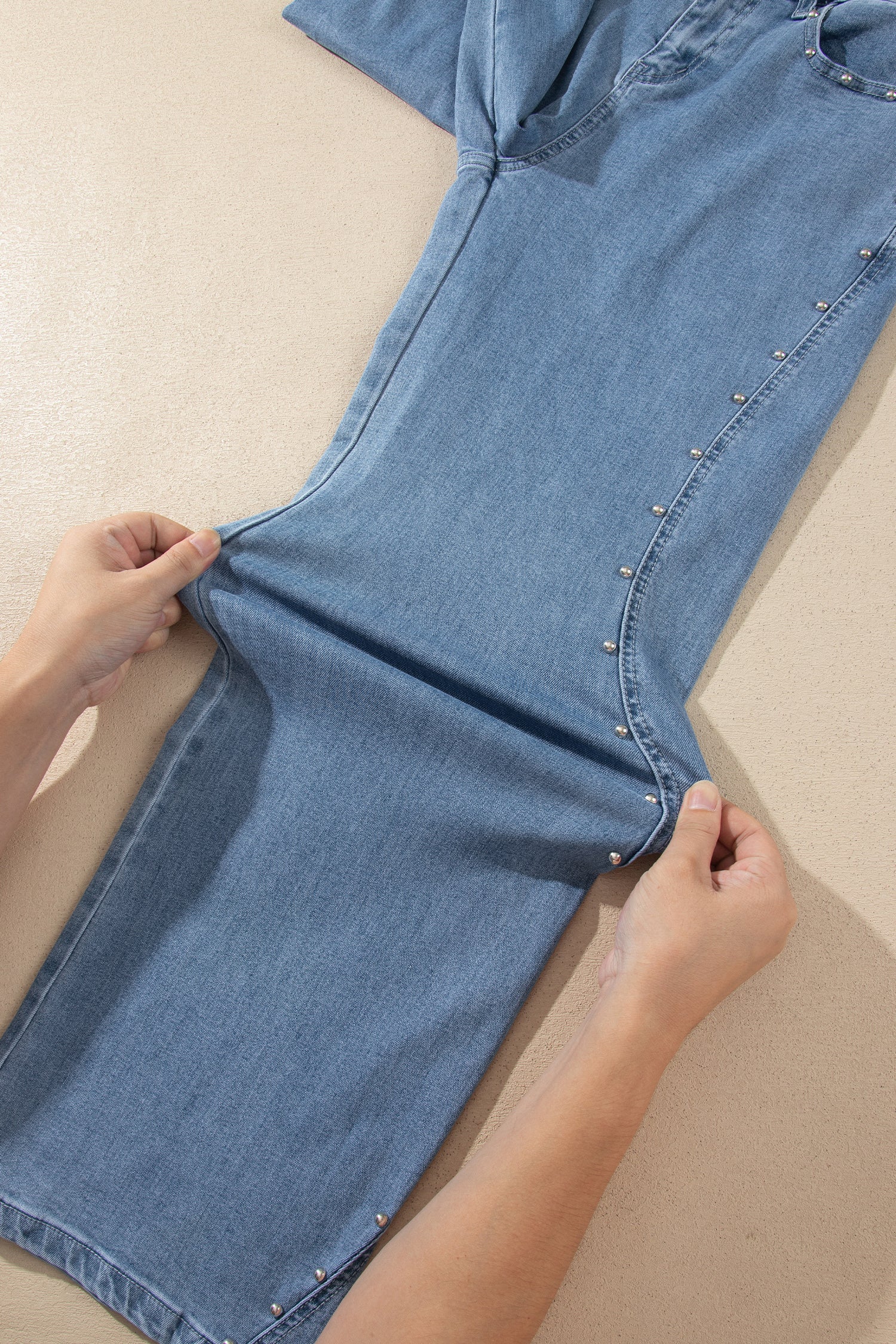 A pair of light blue denim shorts with decorative silver studs along the side, being held and stretched by a person's hands on a beige background.