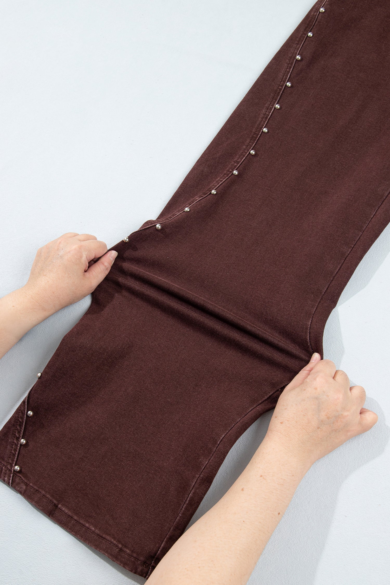 A pair of dark brown pants laid flat on a light gray background, with a person's hands stretching the fabric to show its elasticity. The pants feature decorative silver-colored studs along the seams.