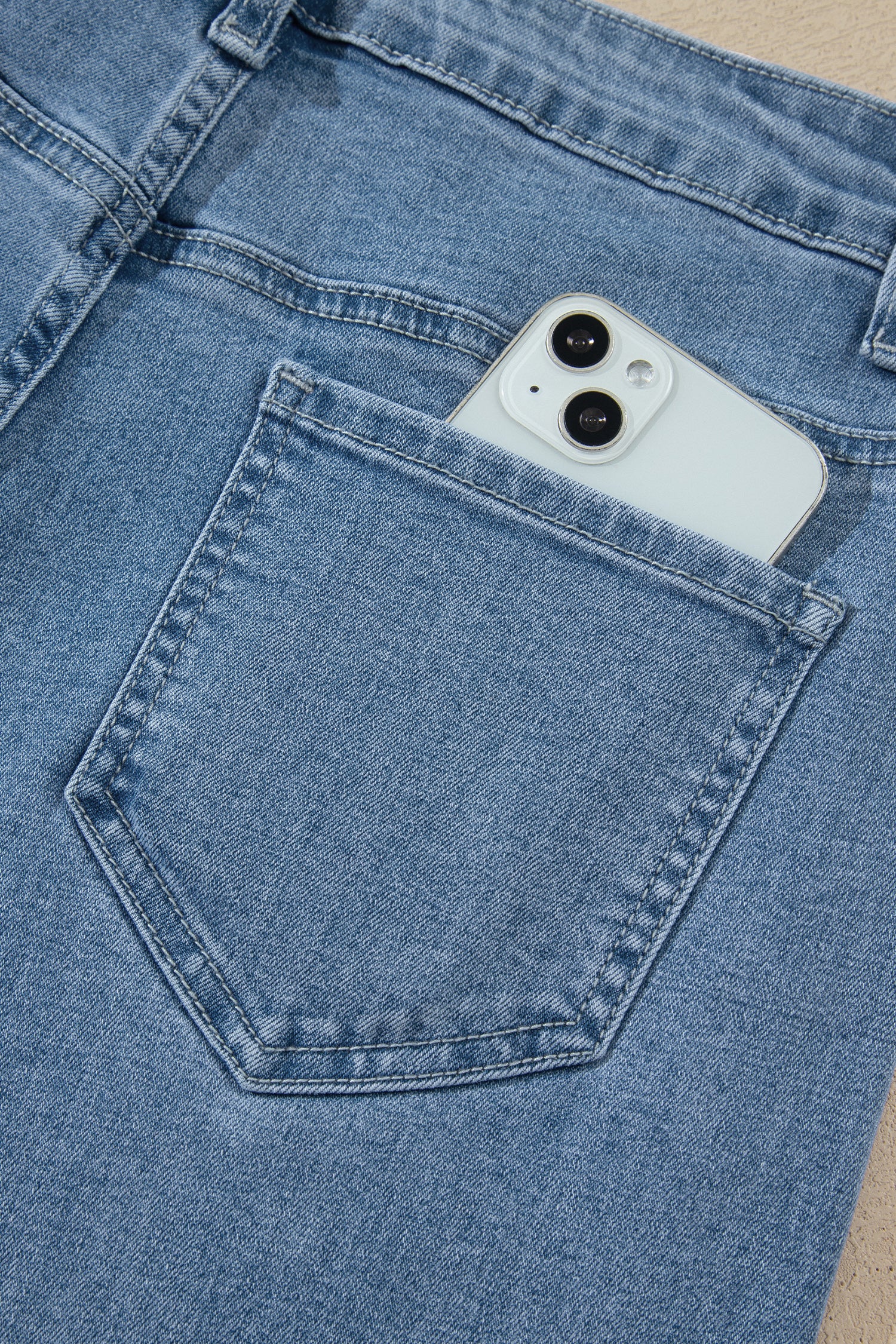 Close-up of a light blue denim pair of jeans with a white smartphone tucked into the back pocket, set against a neutral beige background.