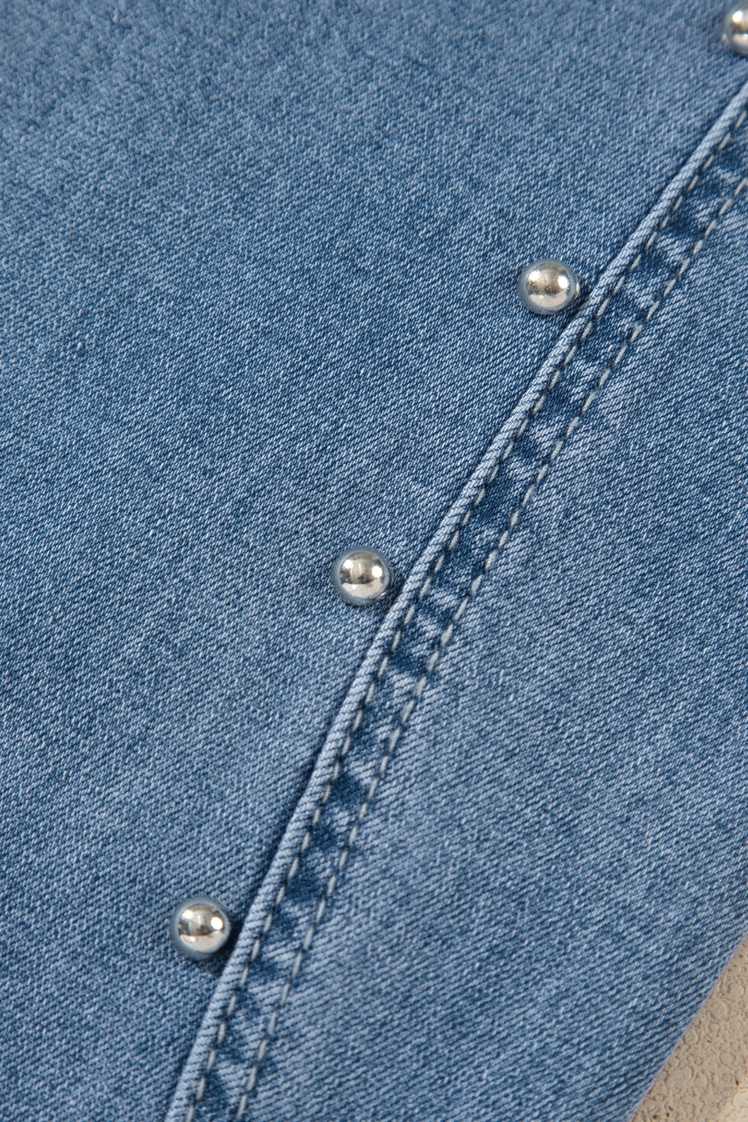 Close-up of a light blue denim fabric with diagonal silver metal studs along a stitched seam.