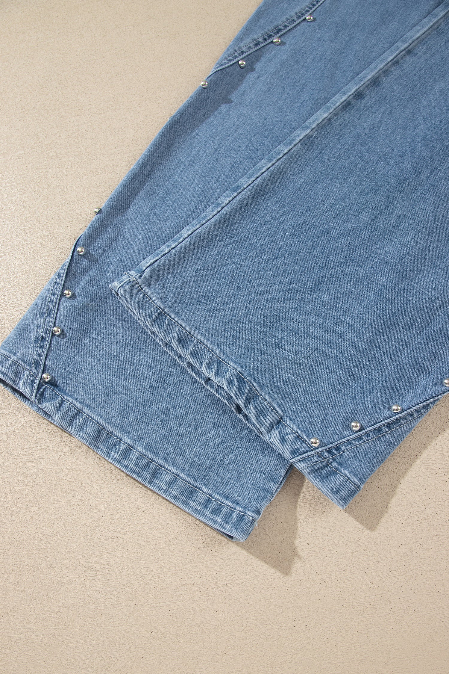 Close-up of a light blue denim garment laid flat on a beige surface, showing the fabric texture, stitching, and small metallic embellishments along the edge.