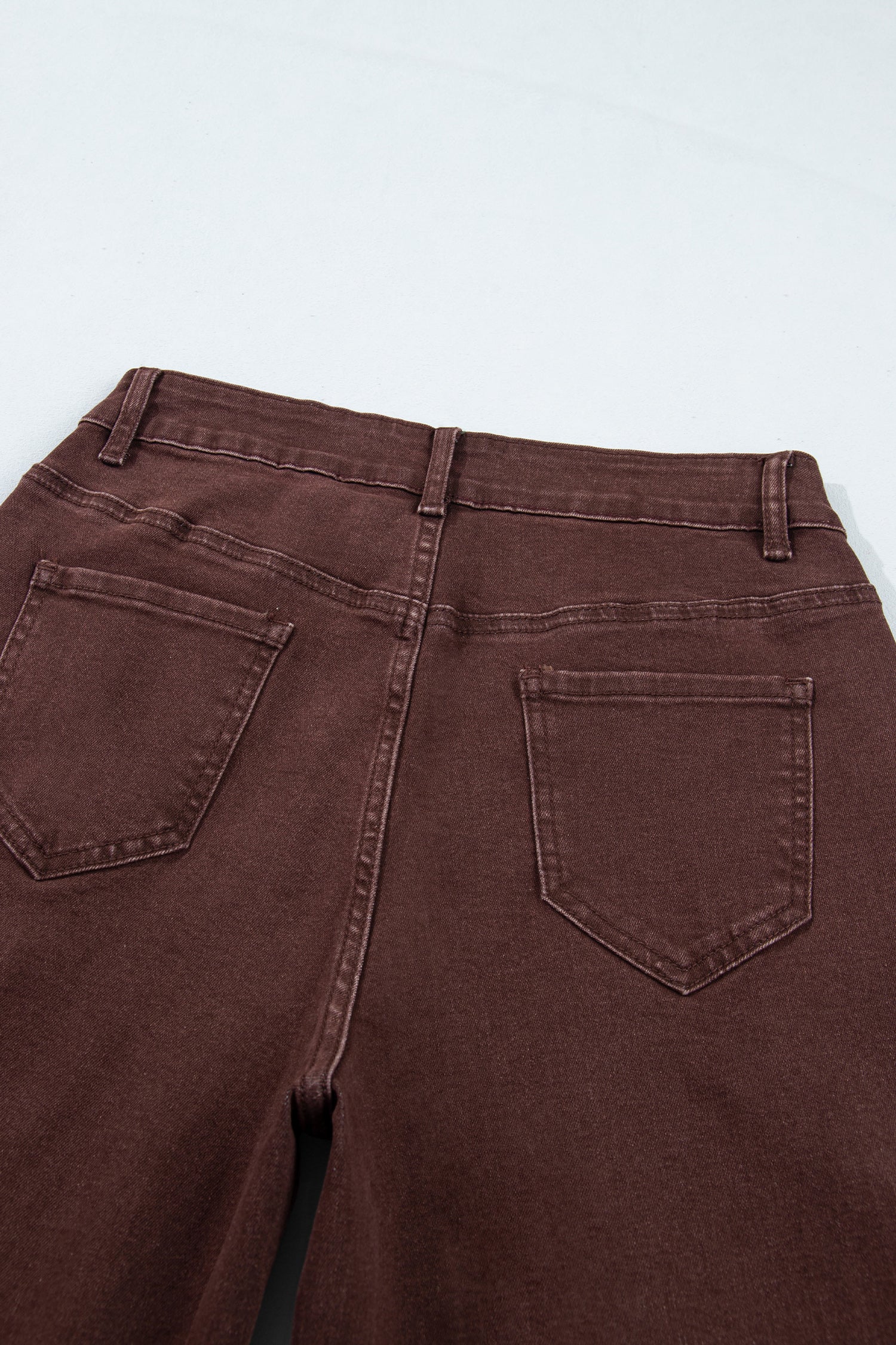 Close-up back view of dark brown denim jeans showing the waistband, belt loops, and back pockets on a light gray background.