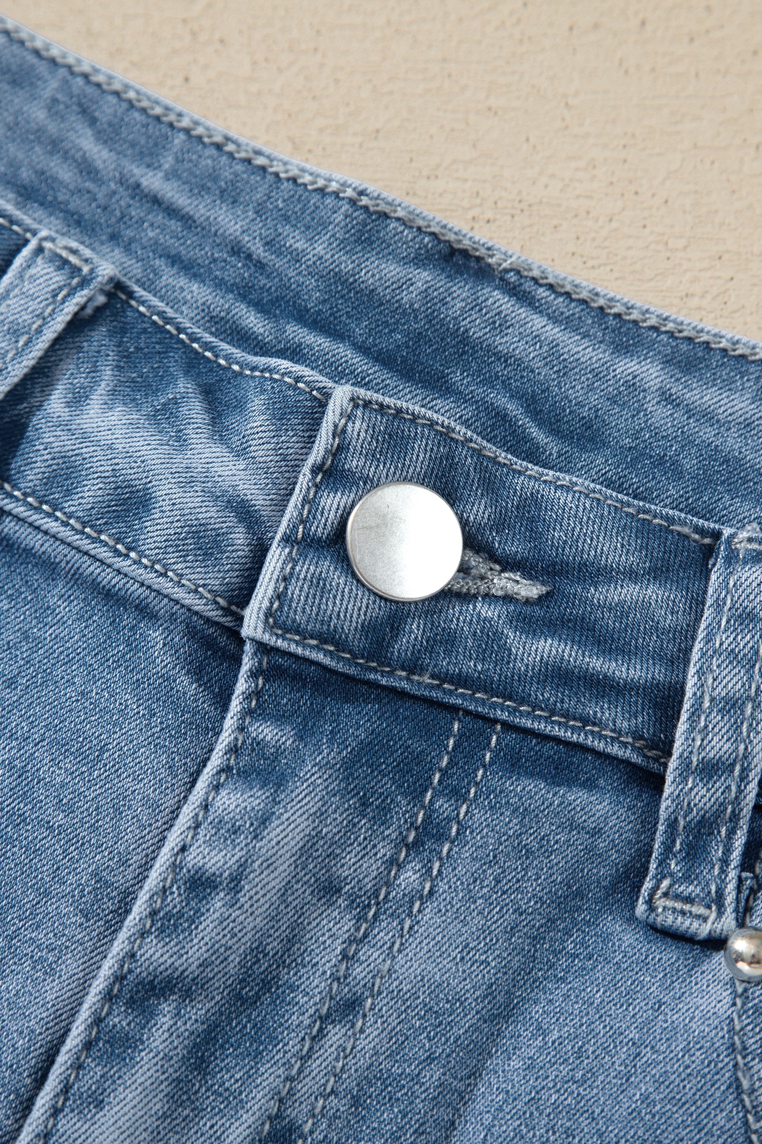 Close-up of blue denim jeans showing the waistband, belt loop, and a silver metal button closure against a light beige background.