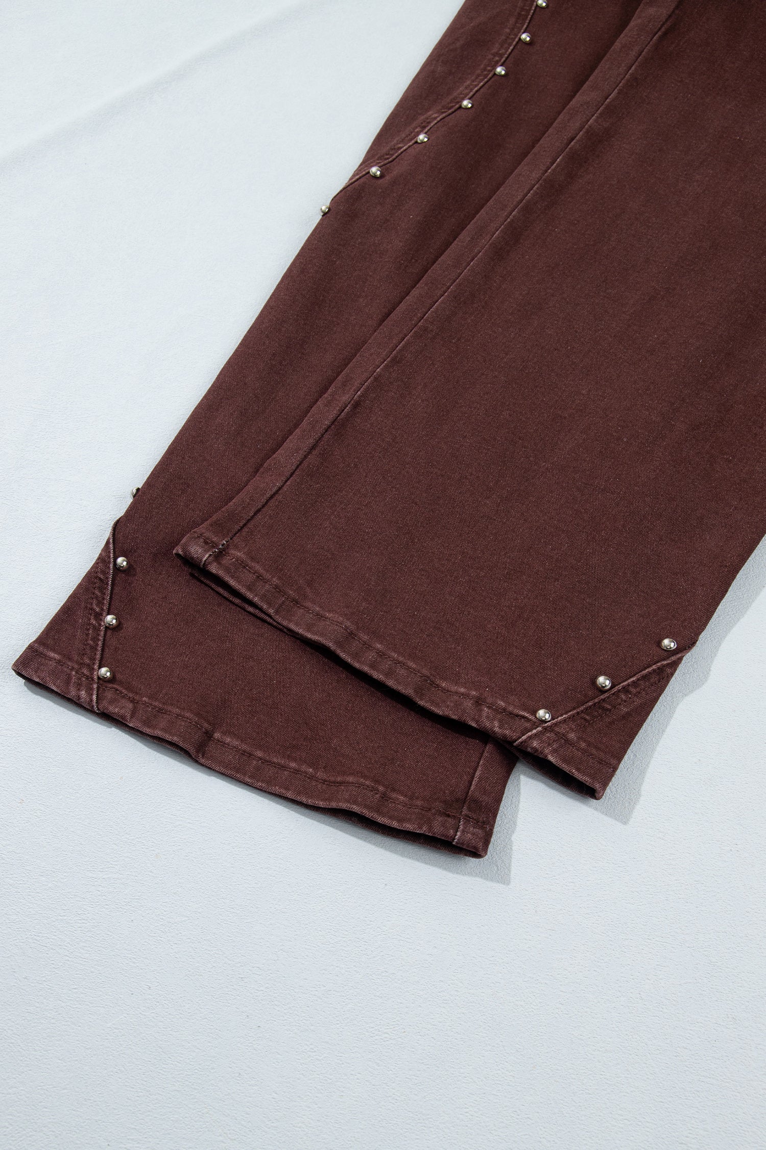 Close-up of a brown fabric garment, likely a pair of pants or jeans, folded over to show the hem and side seam details with small metallic rivet accents on a light gray background.