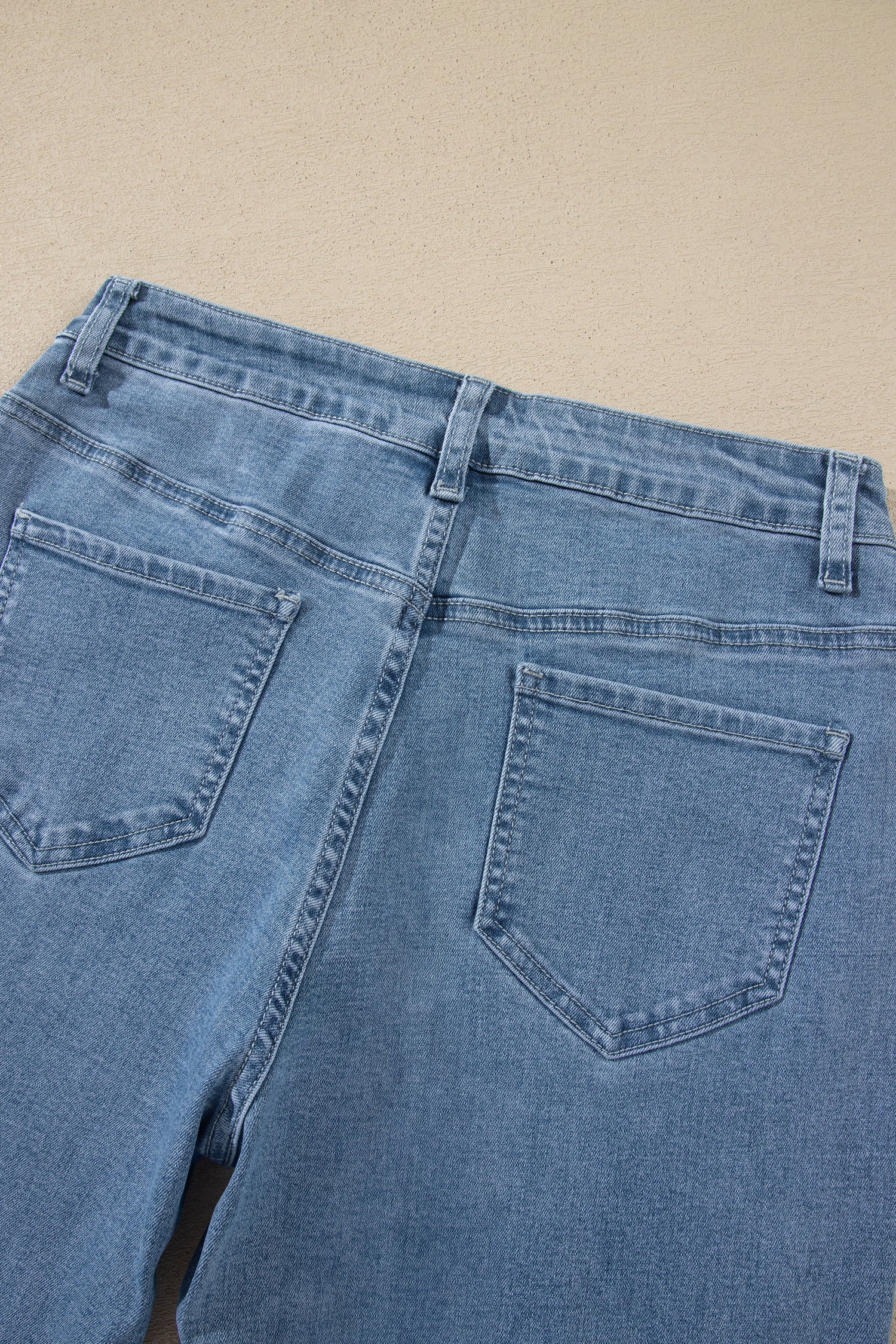 Close-up of the back waistband and pockets of light blue denim jeans laid flat on a beige surface.
