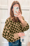 A woman takes a mirror selfie wearing a brown and olive green checkered knit sweater, blue jeans, and holding a clear plastic cup.