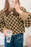 A woman takes a mirror selfie wearing a brown and dark green checkered knit sweater, blue jeans, and layered gold necklaces while holding a clear plastic cup.