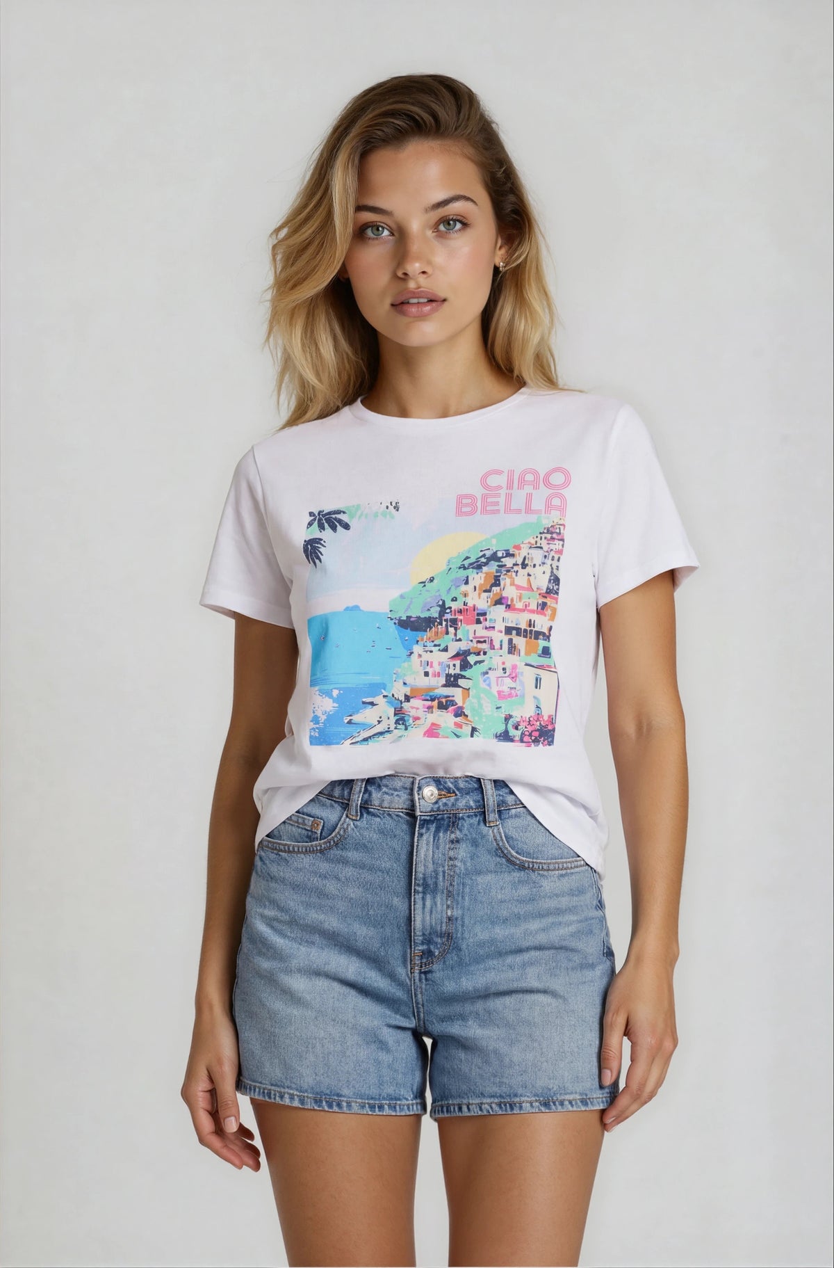Woman wearing a white t-shirt with colorful graphic and 'Ciao Bella' text, paired with denim shorts on a plain background.