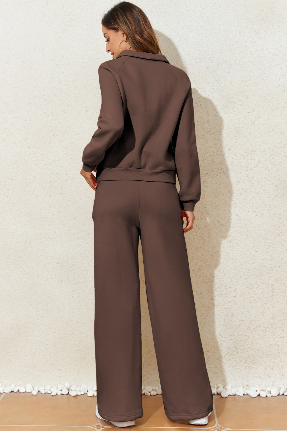 Woman wearing a brown tracksuit against a textured white wall.