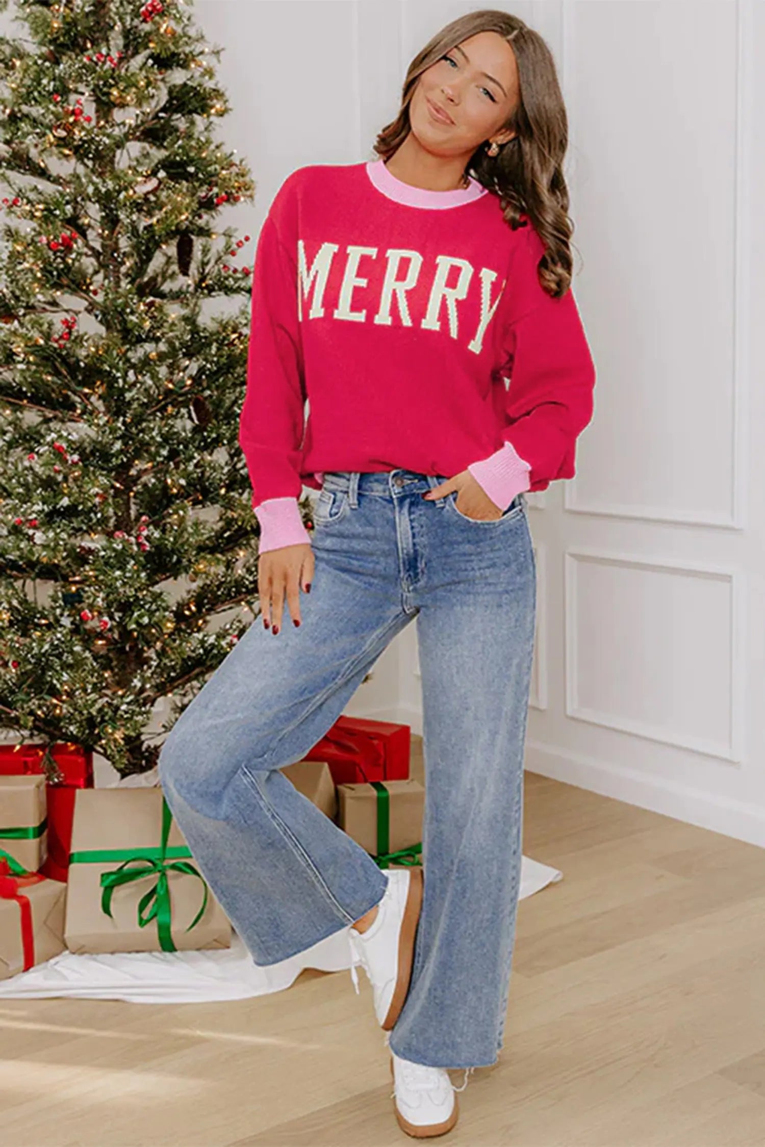 A woman wearing a bright pink crewneck sweatshirt with 'MERRY' printed in white across the chest, paired with light blue wide-leg jeans and white sneakers, standing in a festive room with a decorated Christmas tree and wrapped gifts.