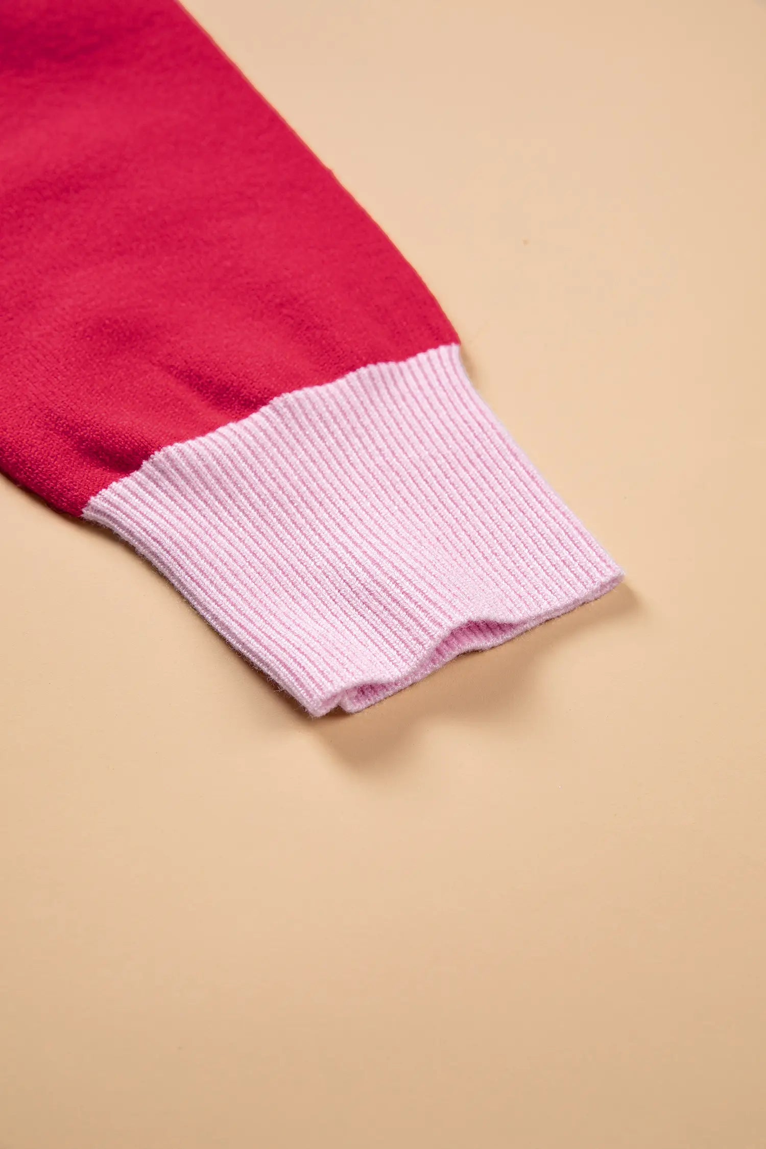 A close-up of a knit garment sleeve showing a solid red upper section and a light pink ribbed cuff, set against a solid beige background.