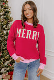 A woman wearing a bright pink crewneck sweatshirt with 'MERRY' printed in white block letters across the chest, paired with light blue jeans. She is standing indoors next to a decorated Christmas tree.