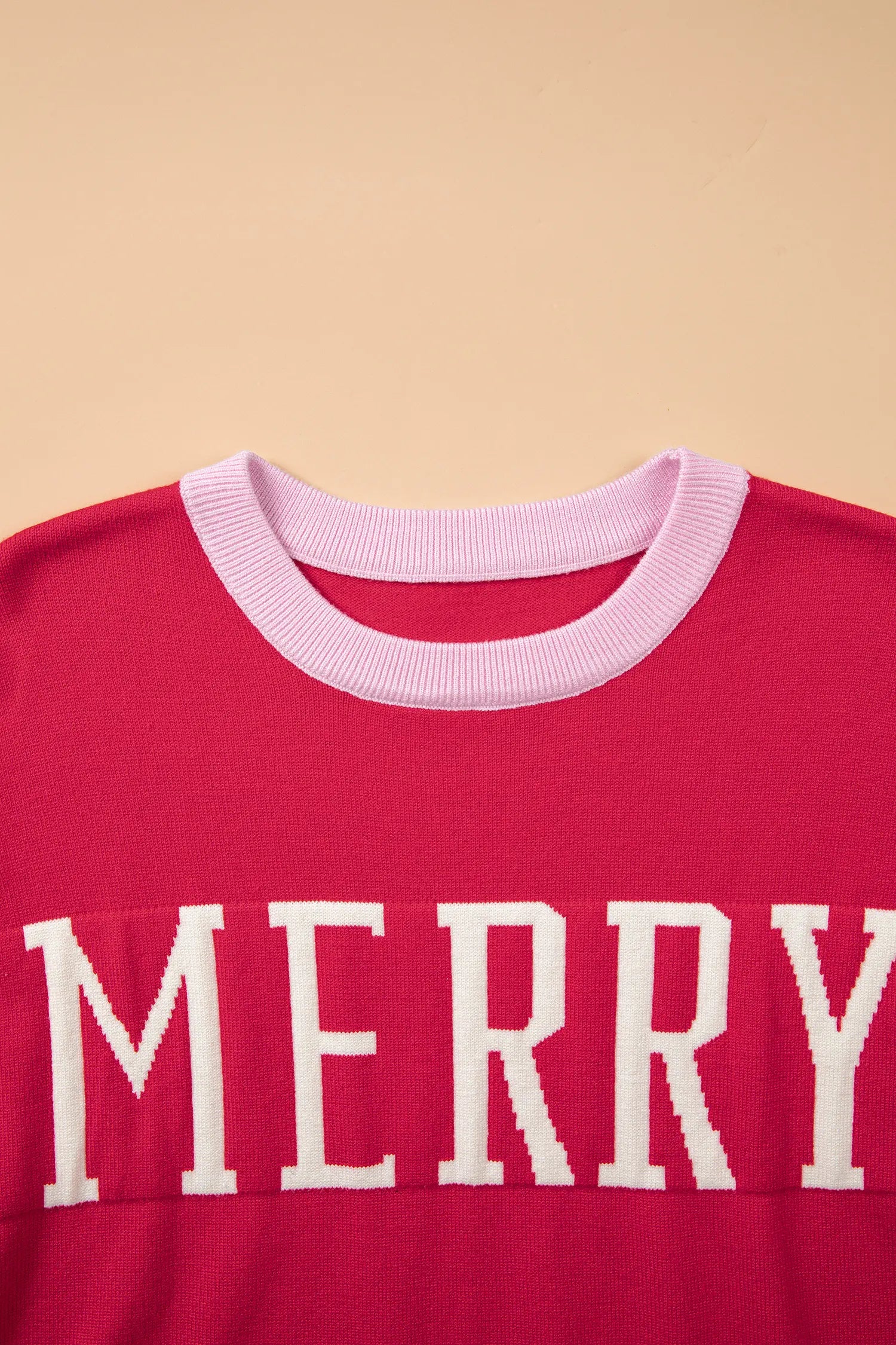 A close-up of a red knit sweater featuring a light pink ribbed crew neck and the word 'MERRY' in large white block letters across the chest, set against a solid beige background.