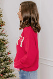 A woman in profile wearing a bright pink crewneck sweater with white knit lettering, standing next to a decorated Christmas tree.