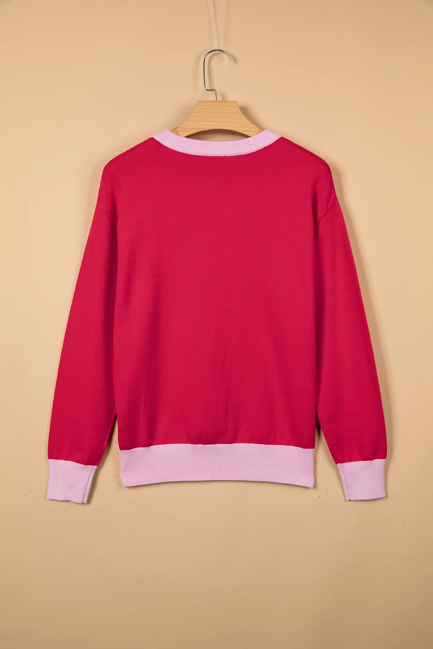 A bright pink long-sleeve sweater with light pink ribbed trim at the crew neck, cuffs, and hem, hanging on a wooden hanger against a solid beige background.