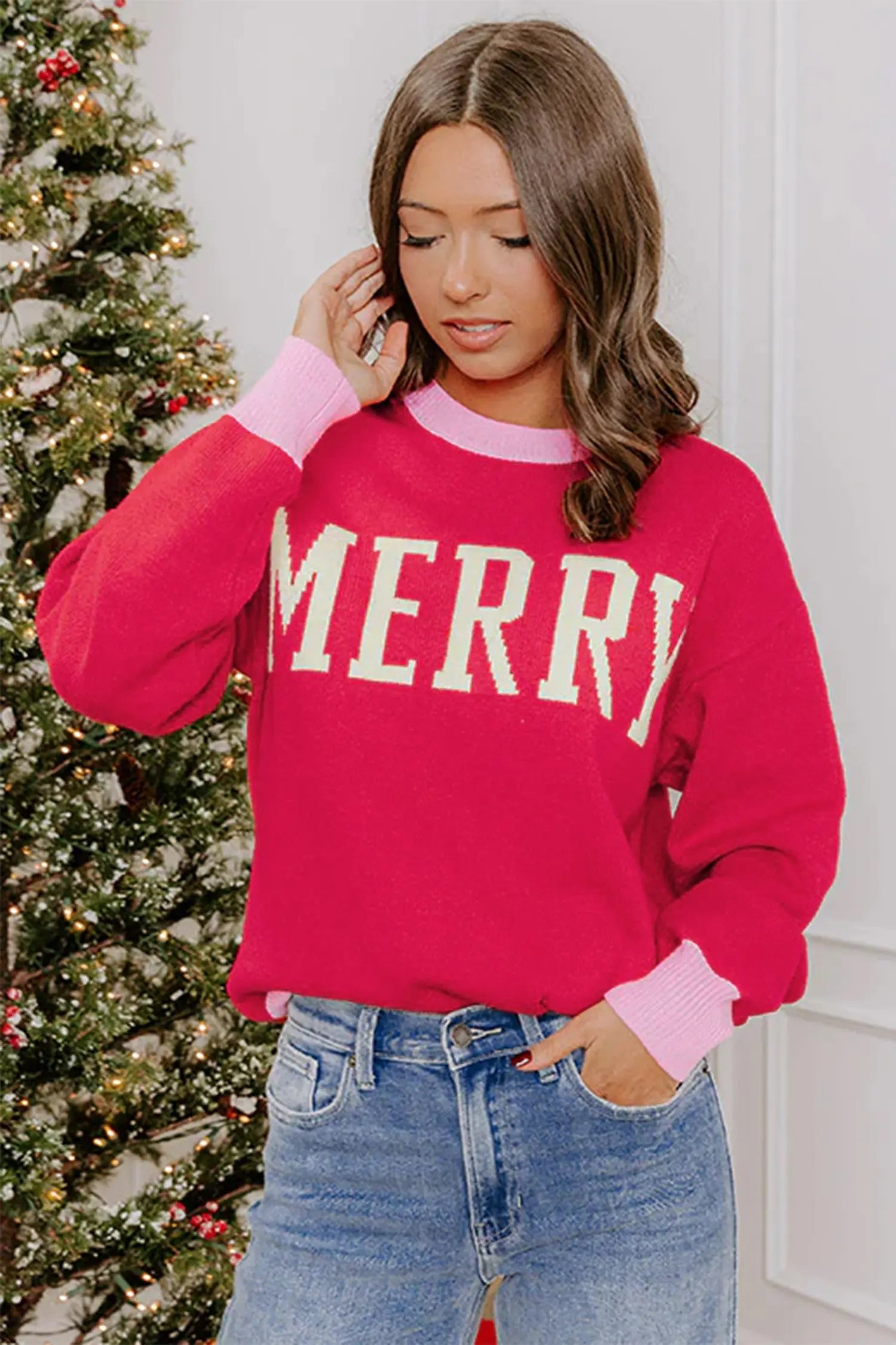 A woman wearing a bright pink crewneck sweater with 'MERRY' in white varsity-style letters across the chest, paired with blue jeans. She is standing indoors next to a decorated Christmas tree.
