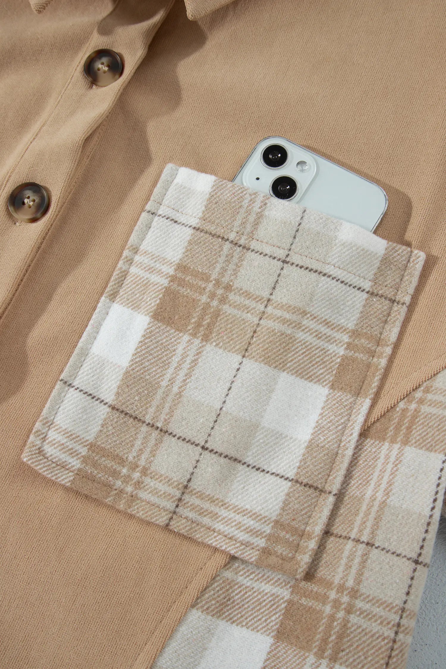 A close-up of a beige button-front shirt pocket holding a white smartphone, with a plaid fabric sleeve or pocket flap layered over the pocket in a tan, cream, and brown check pattern.