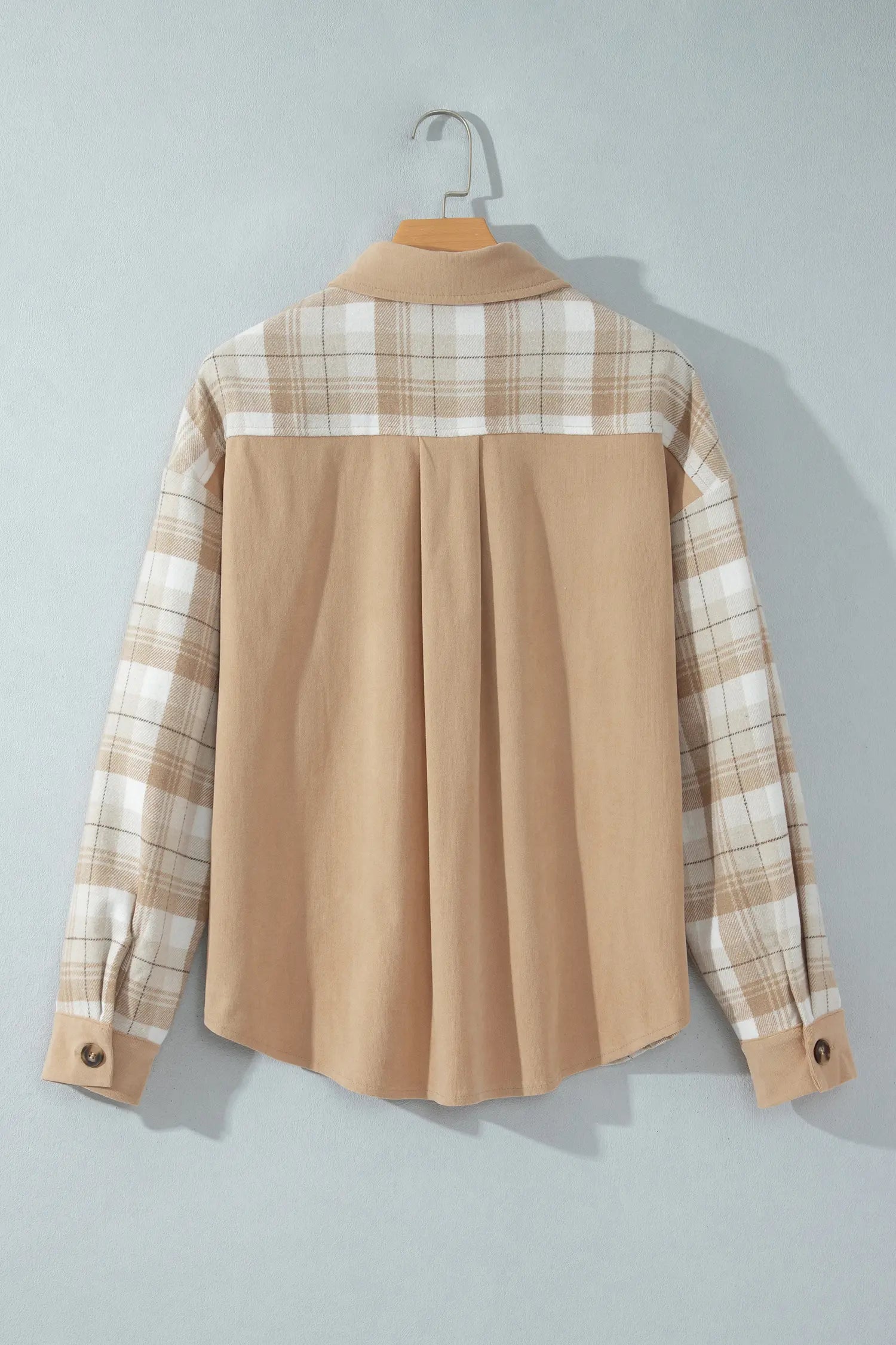 A long-sleeve collared shirt hanging on a wooden hanger against a light gray wall. The back of the shirt is solid tan, while the shoulders and sleeves feature a beige and white plaid pattern.
