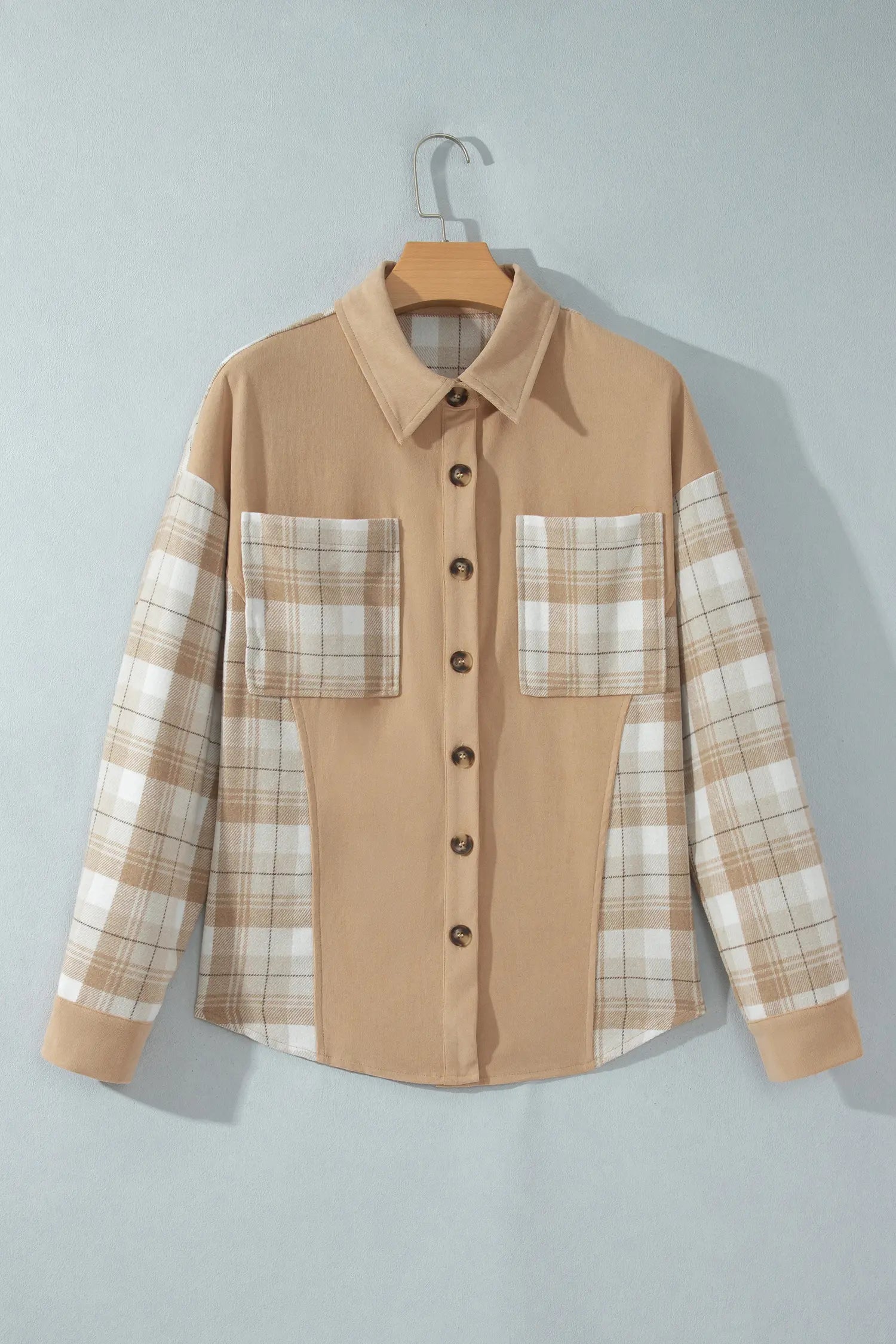 A long-sleeve button-up shirt hanging on a wooden hanger against a light gray wall. The shirt has a solid tan body with beige and white plaid patterned sleeves and matching plaid chest pockets.