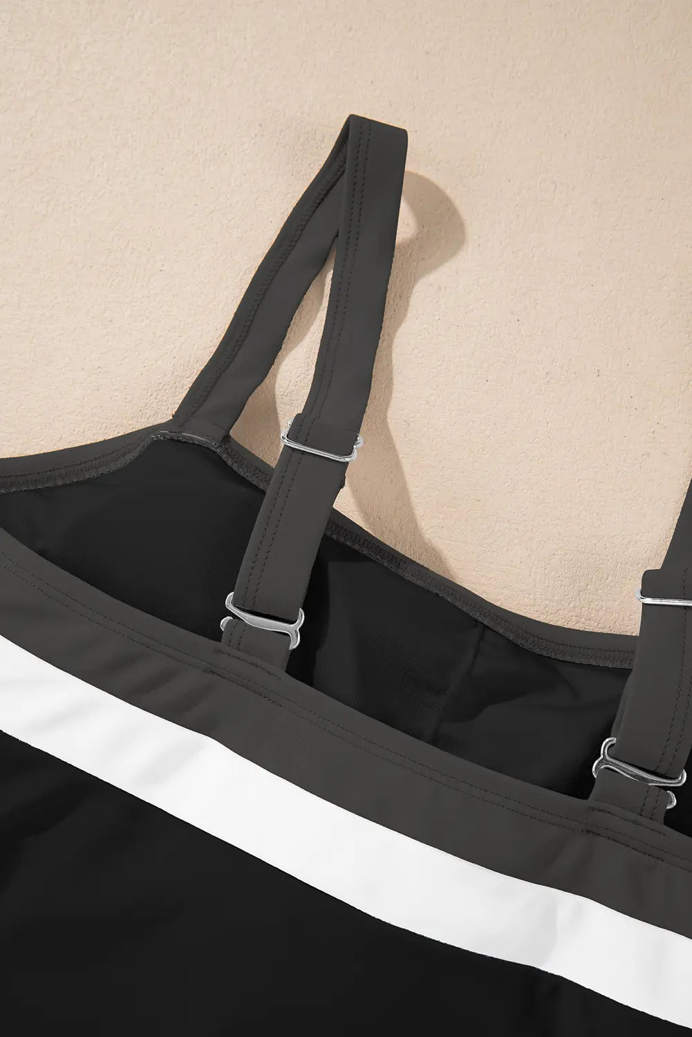Close-up of a black swimsuit or tank top with wide adjustable shoulder straps and a bold white horizontal stripe across the front, shown against a warm beige background.