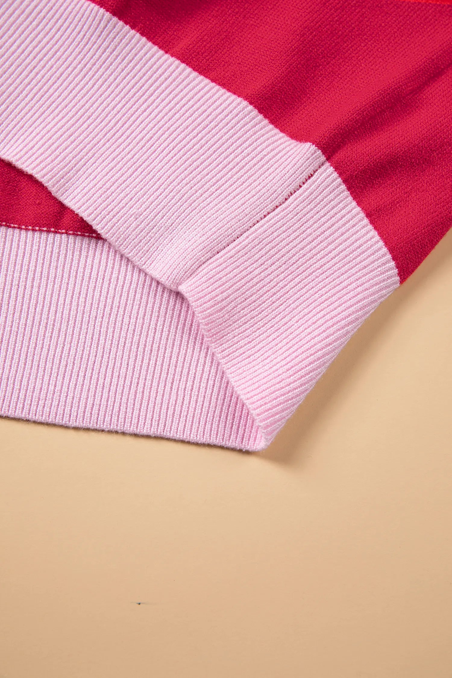 A close-up of a folded garment showing a bright red body with light pink ribbed cuffs, set against a solid beige background.