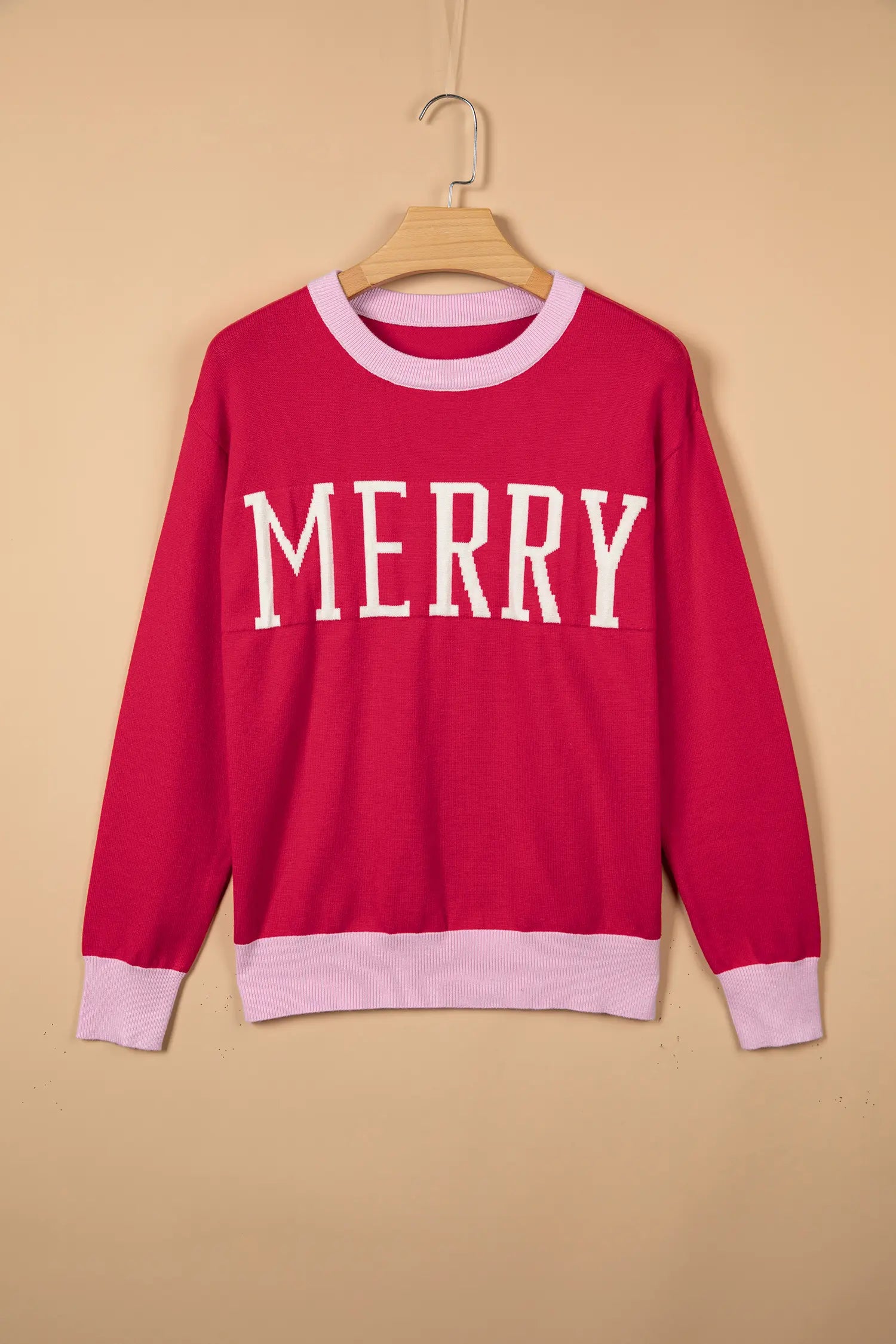 A bright pink crewneck sweater hanging on a wooden hanger against a beige background. The sweater features the word 'MERRY' in white varsity-style letters across the chest, with light pink ribbed trim at the collar, cuffs, and hem.