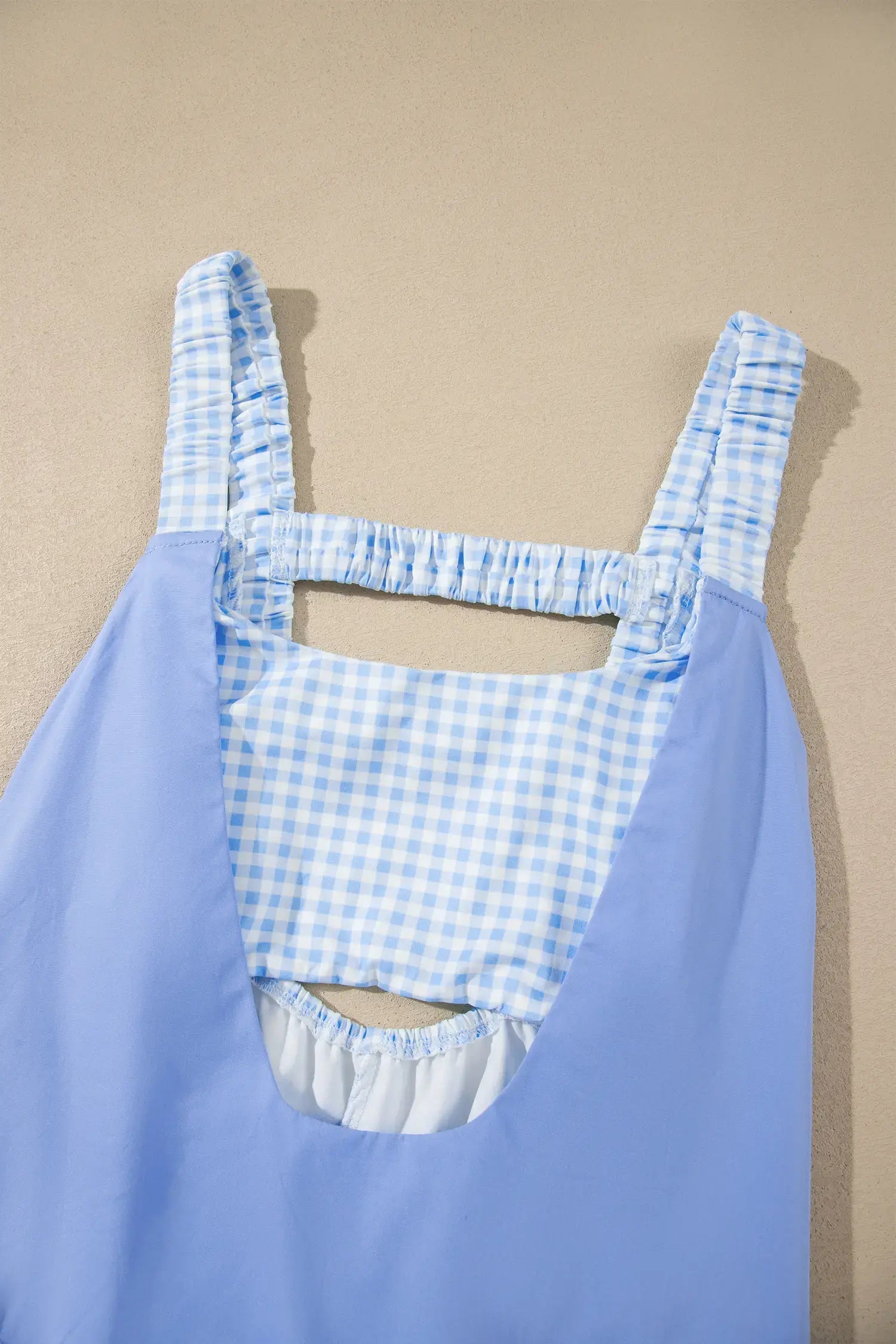 A light blue sleeveless dress with a light blue and white gingham patterned bib front and shoulder straps, shown against a solid beige background.
