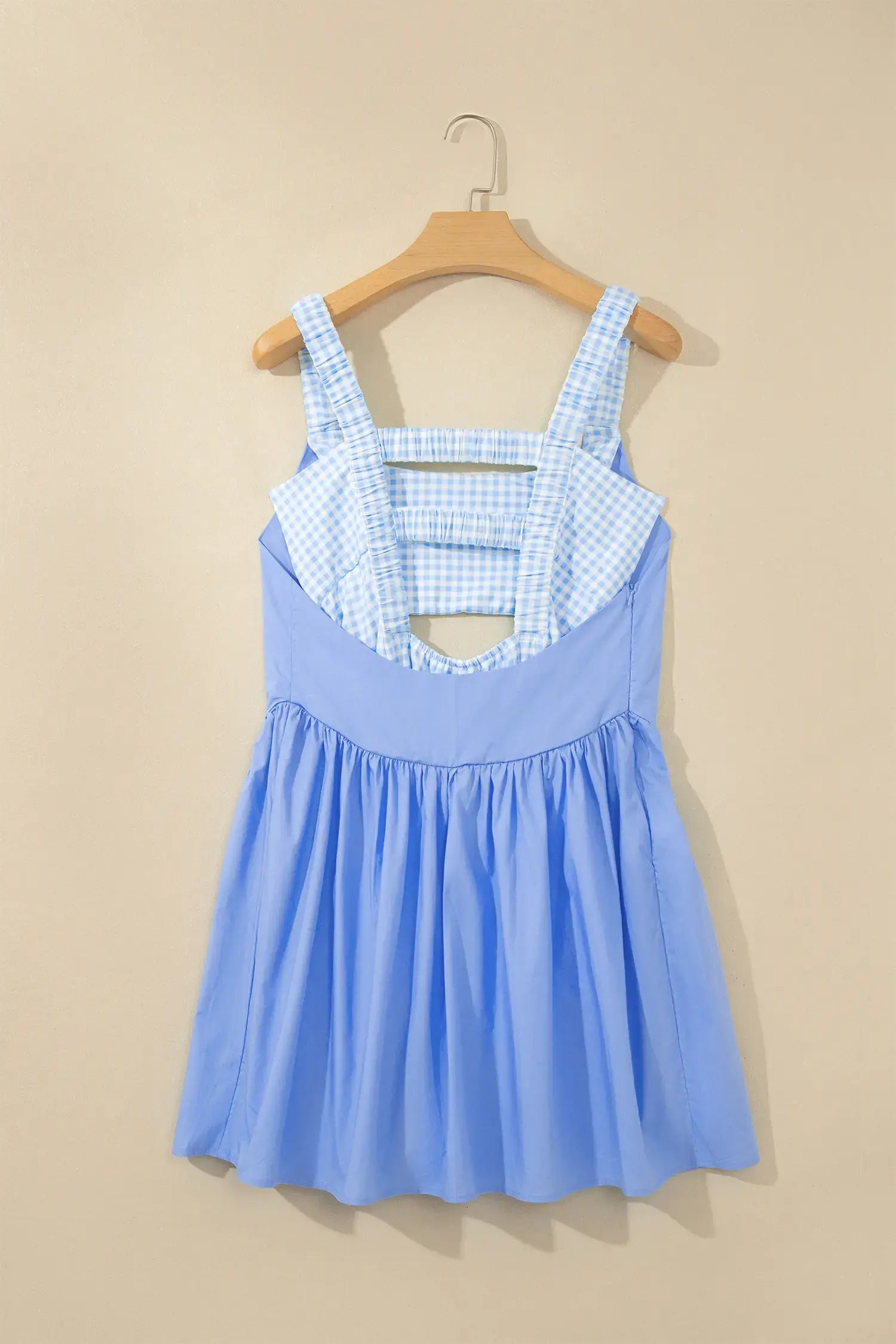 A light blue sleeveless dress with a light blue and white gingham patterned bodice and cutout details, hanging on a wooden hanger against a beige wall.
