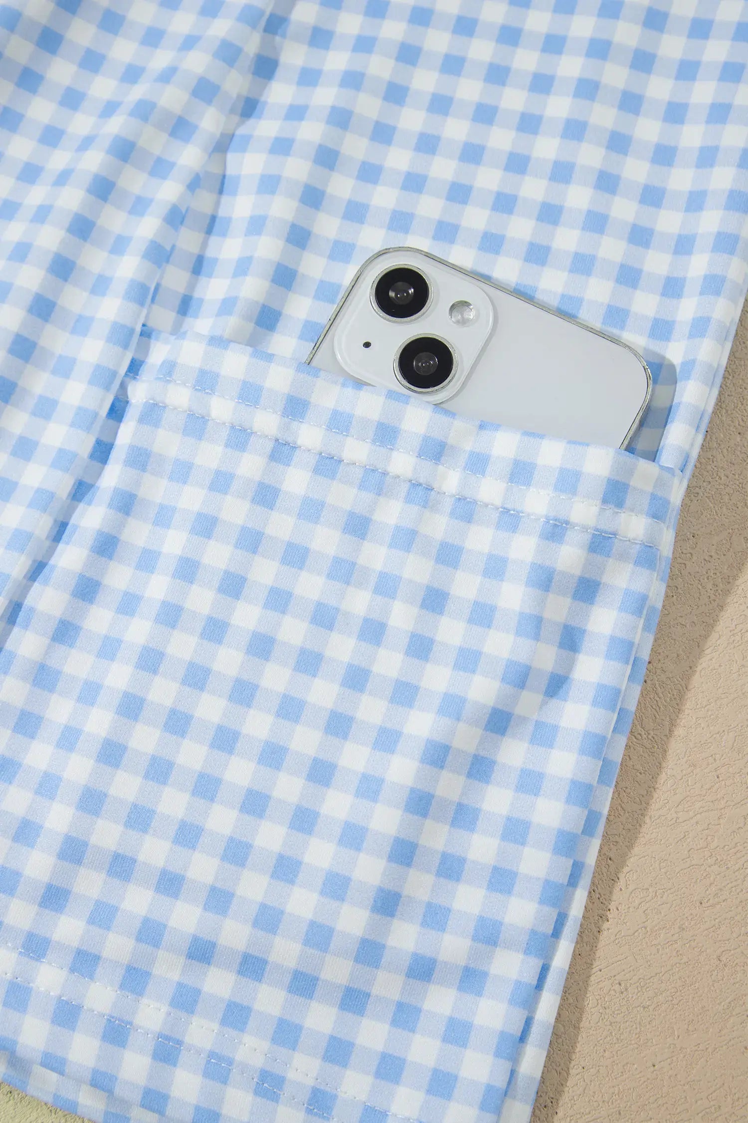 Close-up of a light blue and white gingham pocket with a silver smartphone partially tucked inside, set against a beige textured background.