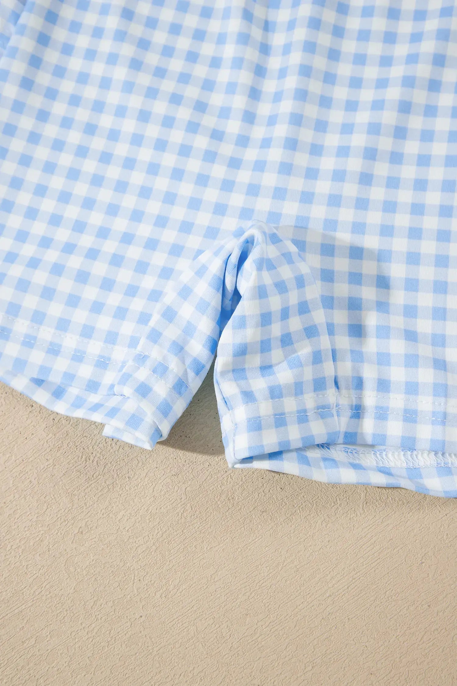 A close-up of a light blue and white gingham fabric, likely a shirt or shorts, folded over to show the texture and pattern against a beige background.