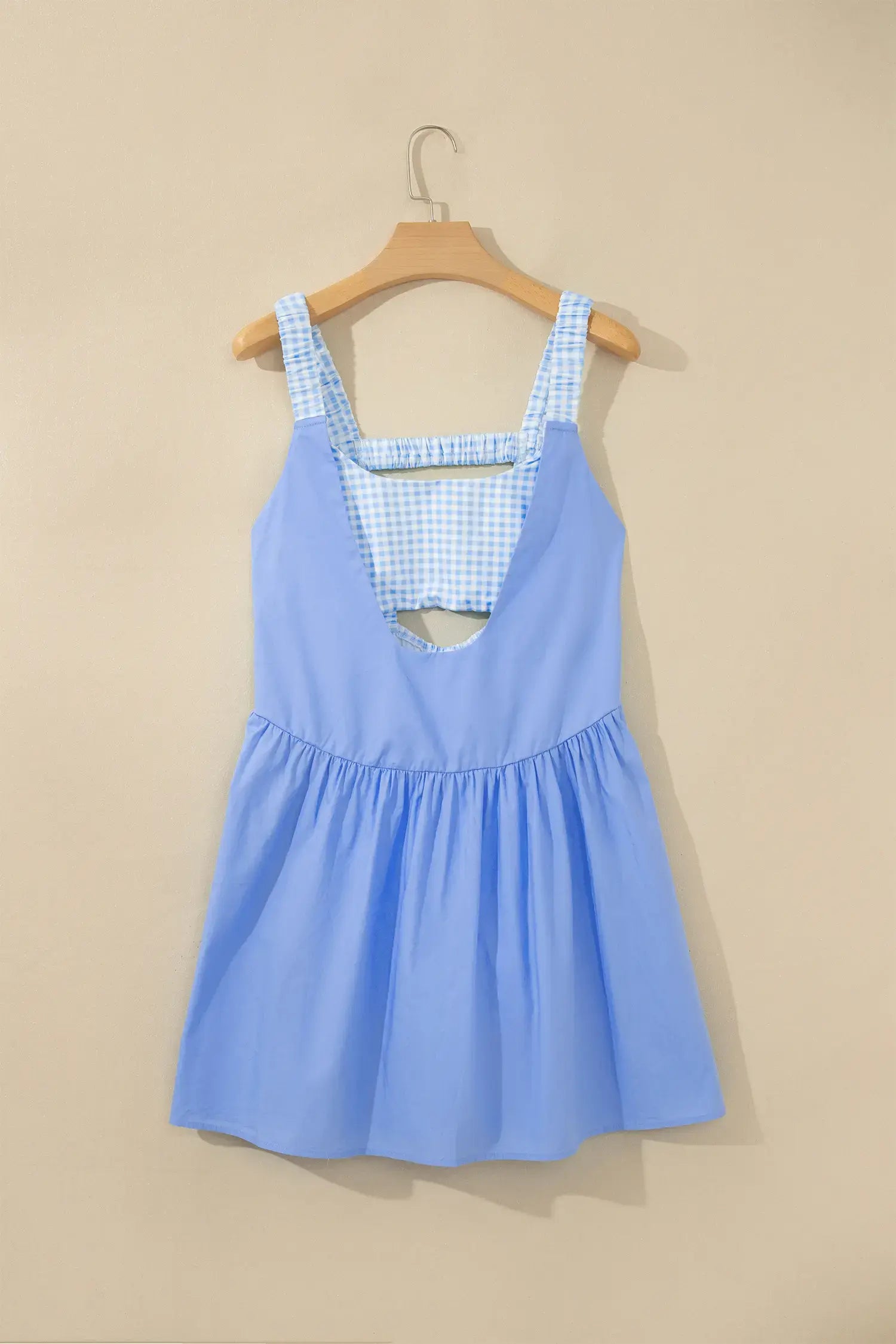 A light blue sleeveless dress with a blue and white gingham patterned neckline and straps, hanging on a wooden hanger against a beige wall.