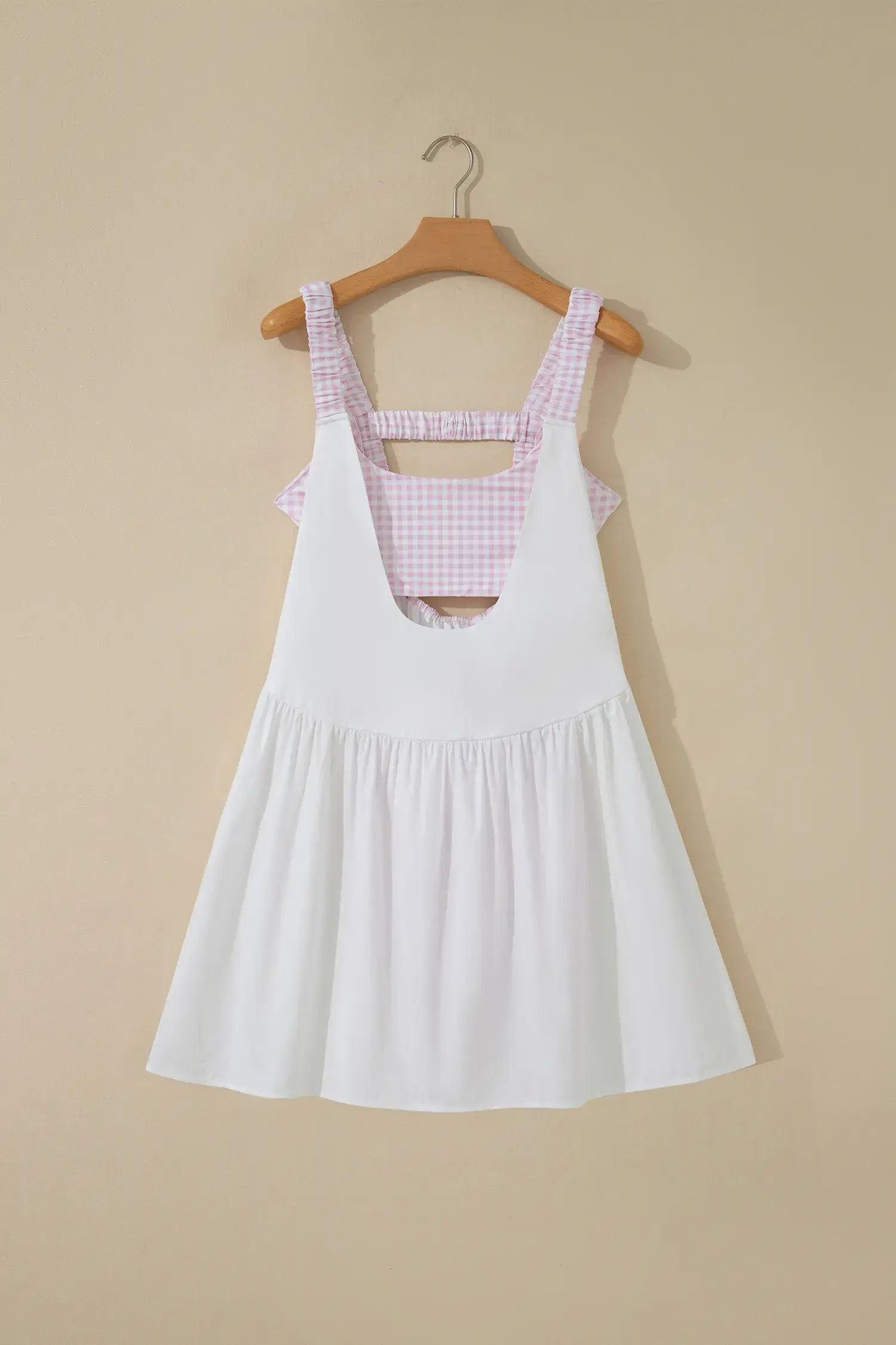 A white sleeveless pinafore-style dress with a pink and white gingham patterned underbust band and shoulder straps, hanging on a wooden hanger against a beige wall.