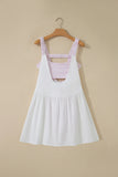 A white sleeveless pinafore-style dress with a pink and white gingham patterned underbust band and shoulder straps, hanging on a wooden hanger against a beige wall.