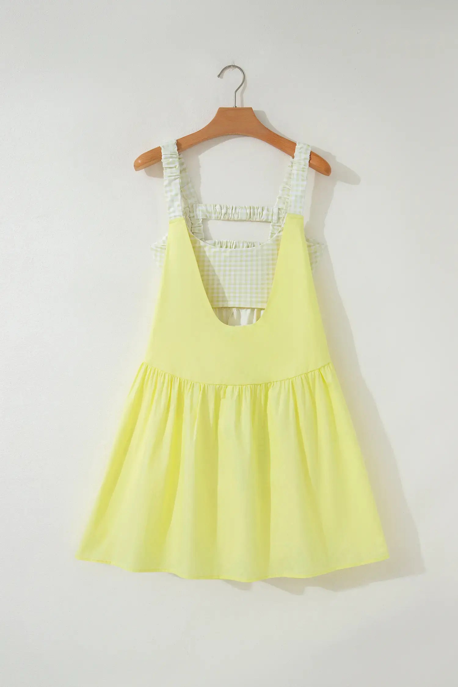 A light yellow sleeveless pinafore-style dress with a white and green gingham patterned inner bodice and shoulder straps, hanging on a wooden hanger against a plain off-white wall.