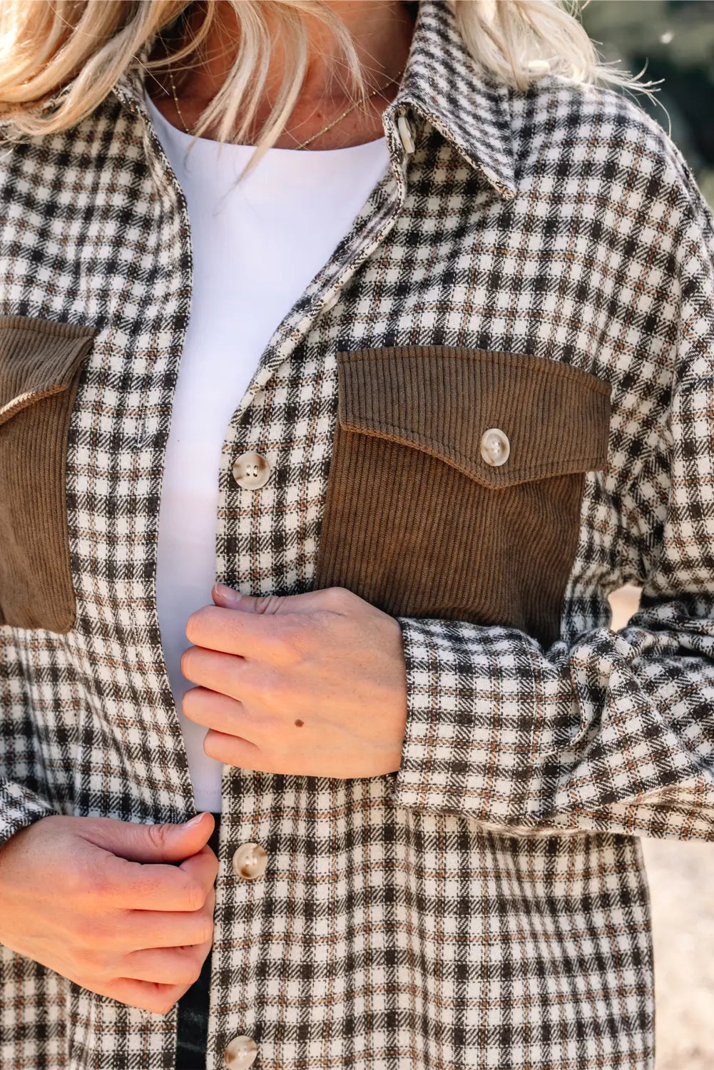 A close-up of a person wearing a brown and beige plaid button-up shirt with brown corduroy chest pockets over a white top.