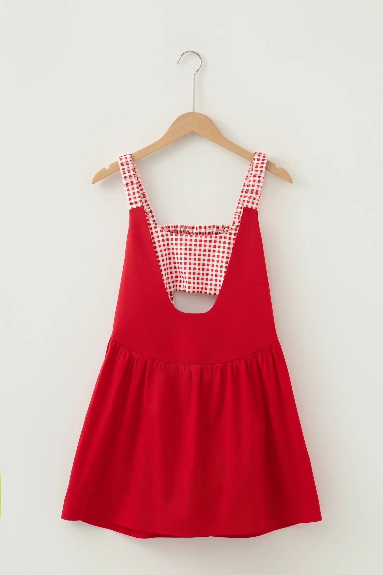 A red sleeveless babydoll dress with a red and white gingham patterned bib and shoulder straps, hanging on a wooden hanger against a plain off-white wall.