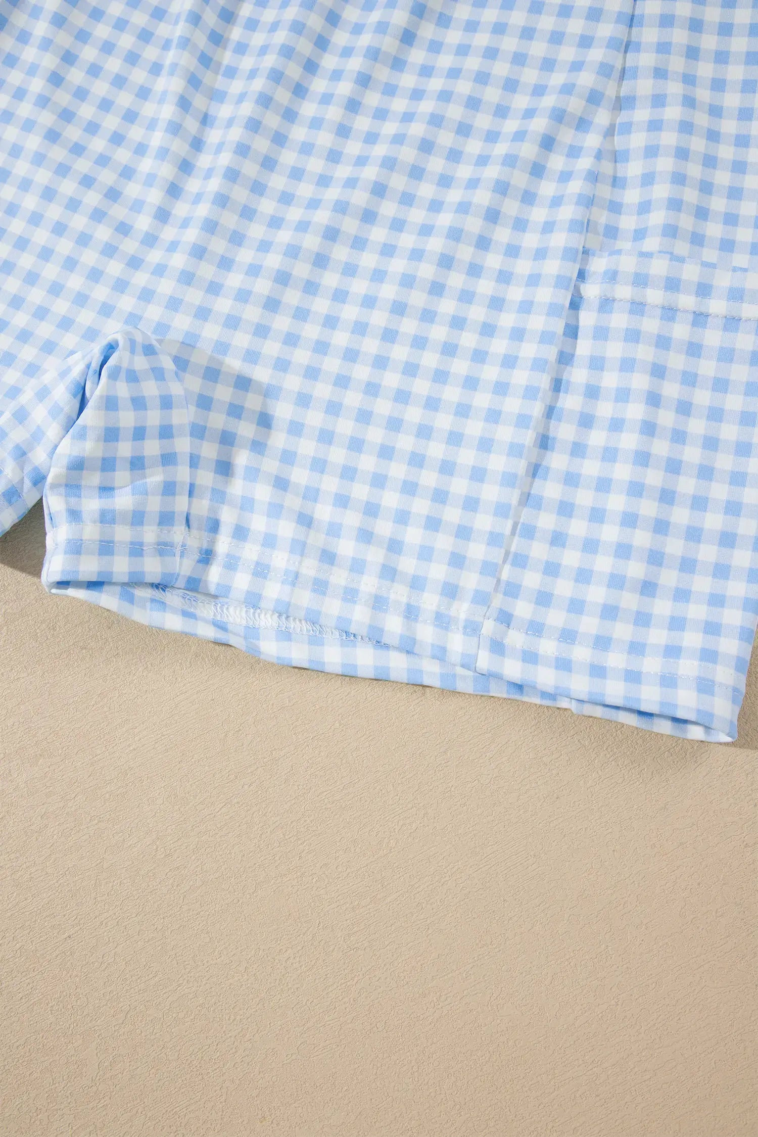 A close-up of a light blue and white gingham fabric, likely a shirt or shorts, laid flat on a beige surface.