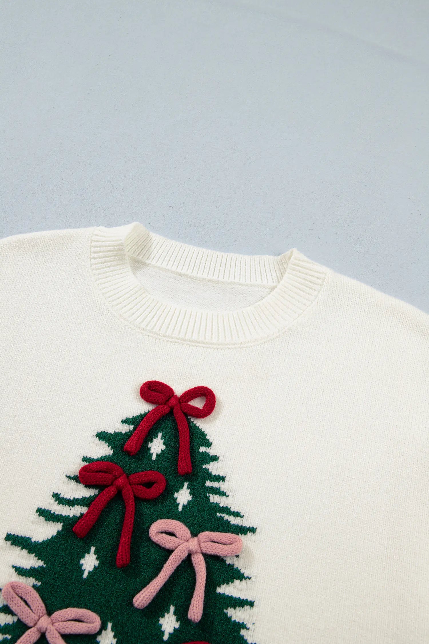 Close-up of a cream knit sweater with a festive Christmas tree, red and pink bows, and white stars.