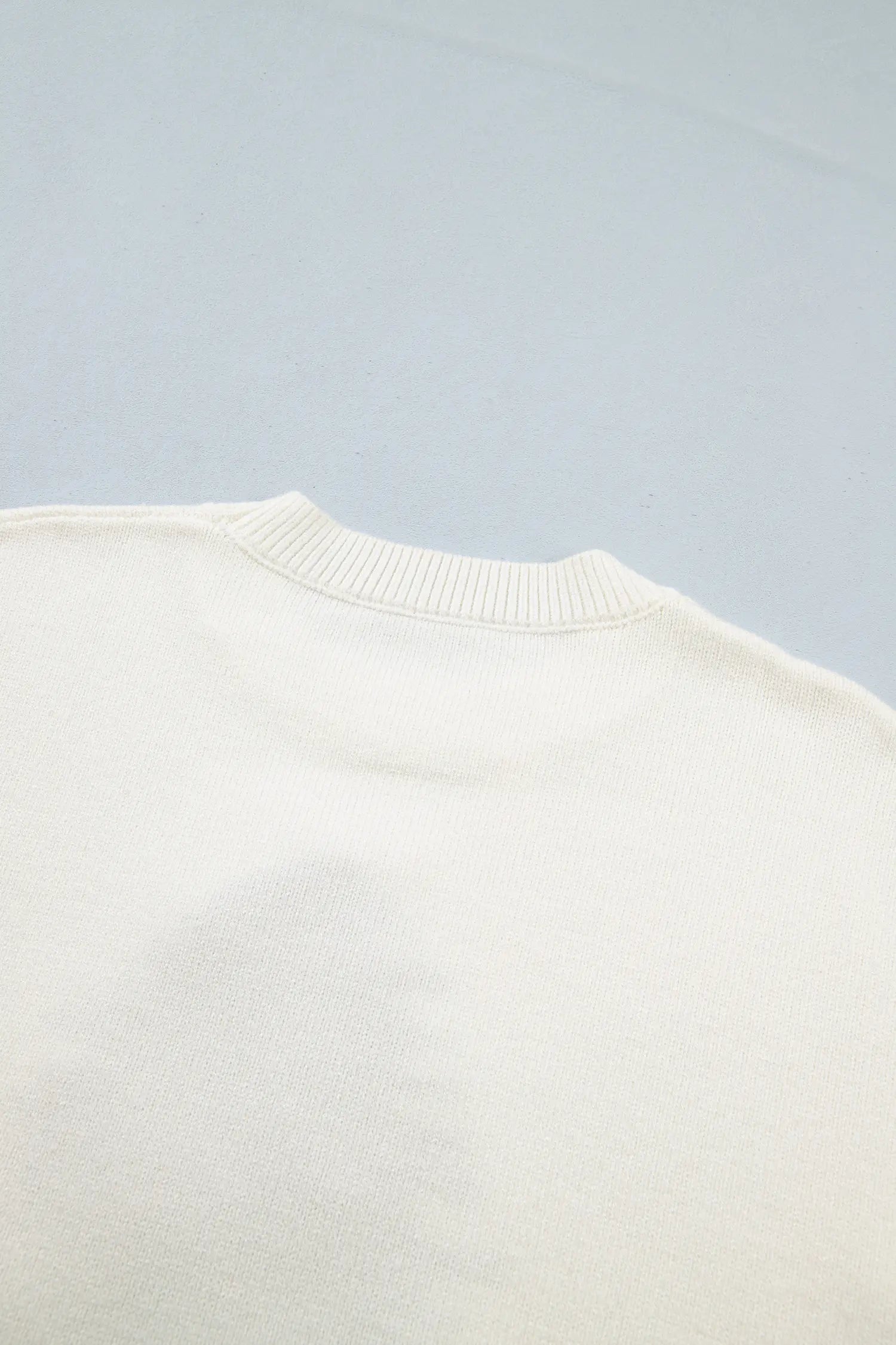 Close-up of a cream knit sweater with a ribbed crew neck against a blue-gray background.