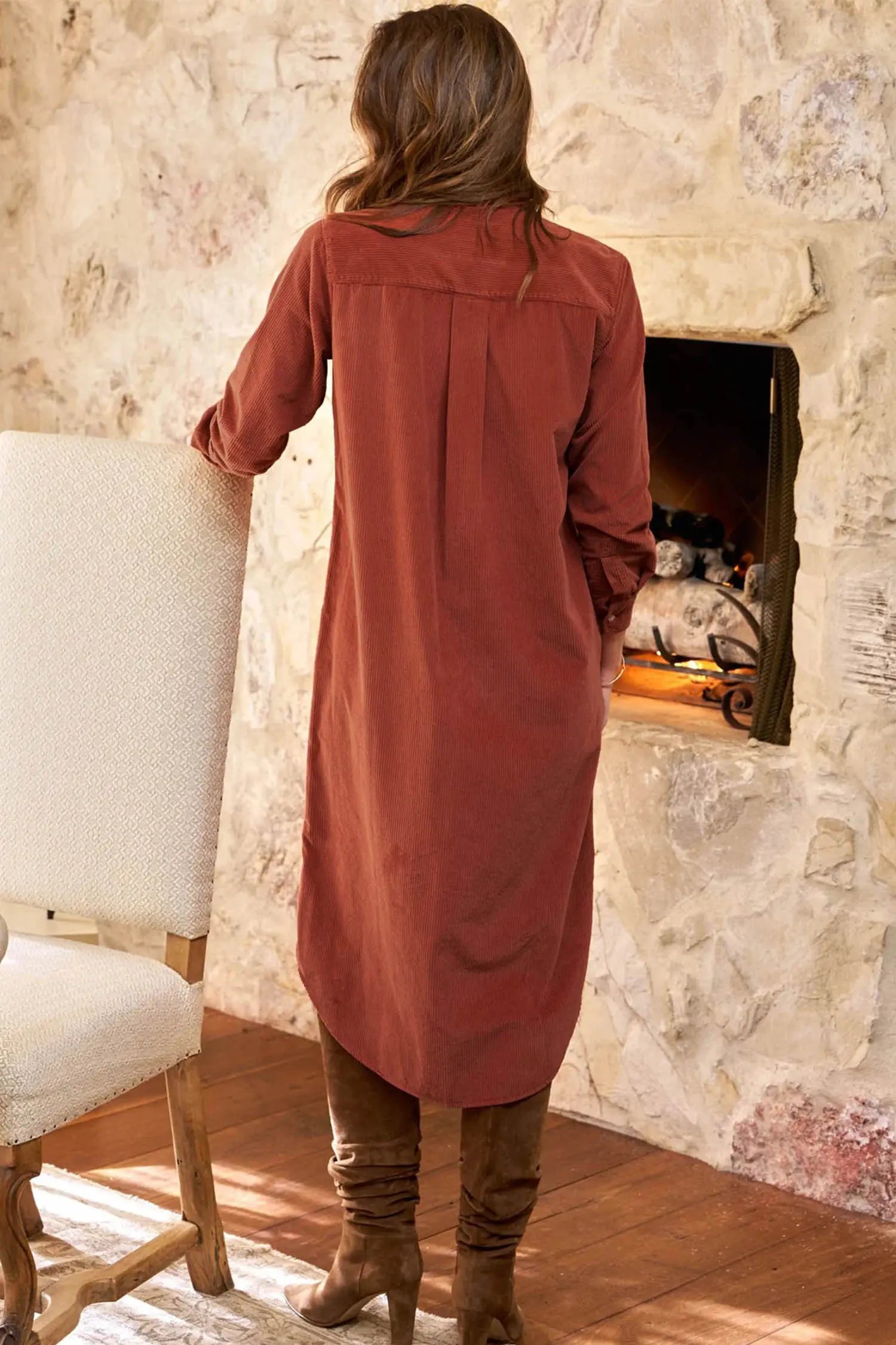 A woman viewed from behind wearing a rust-colored long-sleeve linen tunic dress, standing in front of a stone fireplace in a rustic room.