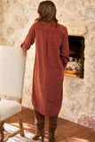 A woman viewed from behind wearing a rust-colored long-sleeve linen tunic dress, standing in front of a stone fireplace in a rustic room.