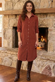 A woman wearing a rust-colored corduroy shirt dress with brown leather knee-high boots, standing in front of a rustic stone fireplace with a fire burning inside.