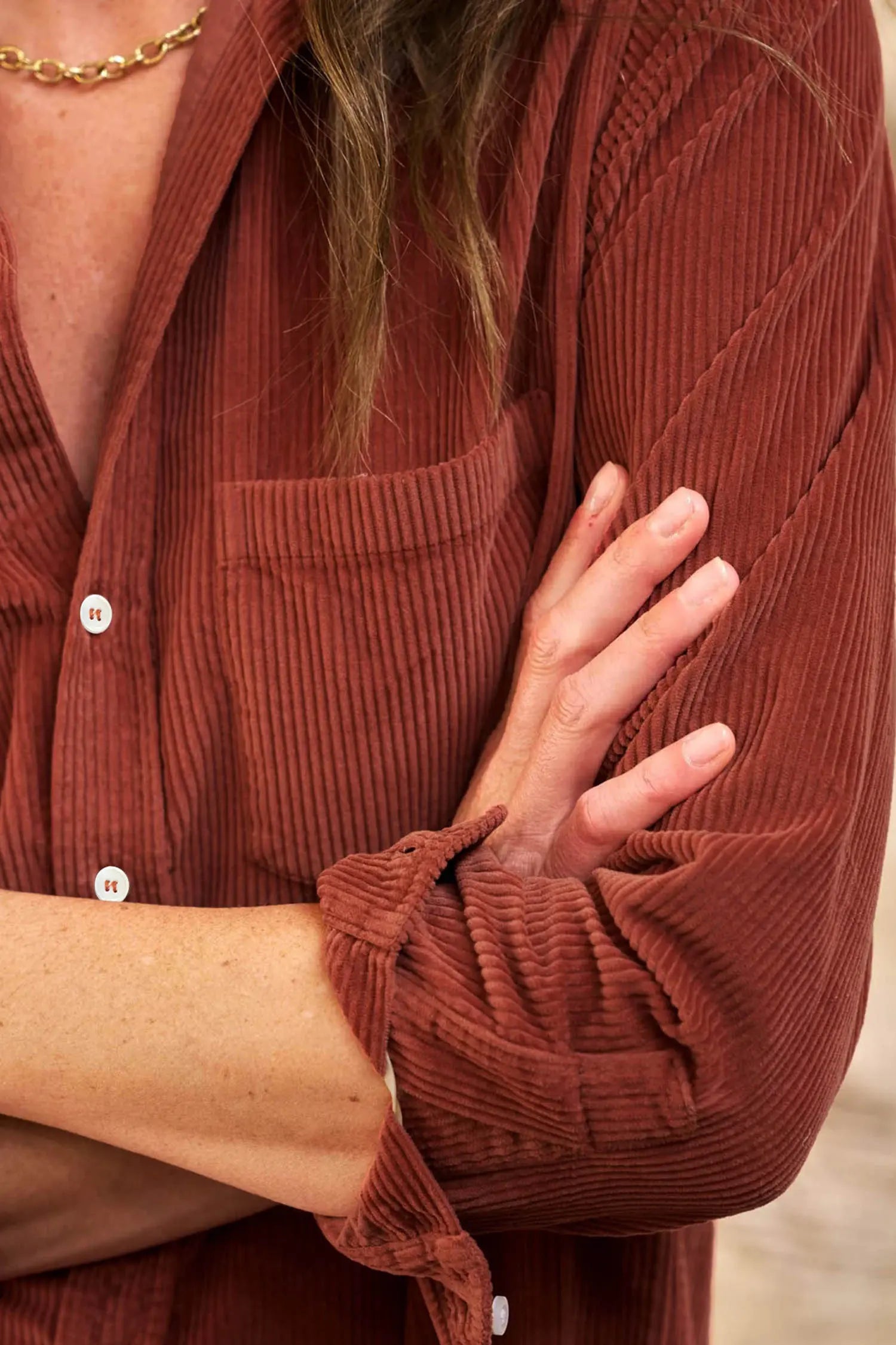 Close-up of a person wearing a rust-brown corduroy button-up shirt with a chest pocket and rolled sleeves, with a gold chain necklace partially visible.