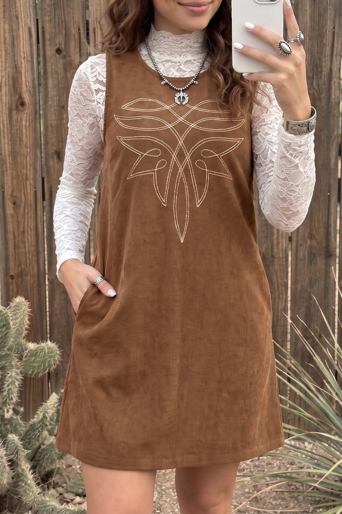 A woman takes a mirror selfie wearing a sleeveless brown corduroy dress with a white embroidered geometric pattern on the chest, layered over a white lace long-sleeve top. The background is a rustic wooden fence with some desert plants.