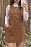 A woman takes a mirror selfie wearing a sleeveless brown corduroy dress with a white embroidered geometric pattern on the chest, layered over a white lace long-sleeve top. The background is a rustic wooden fence with some desert plants.