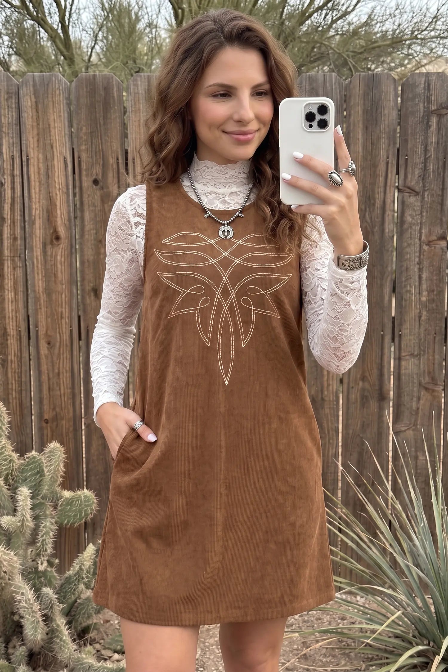 A woman takes a mirror selfie wearing a brown corduroy sleeveless dress over a white lace long-sleeve turtleneck. The dress features a white embroidered geometric design on the chest and side pockets.